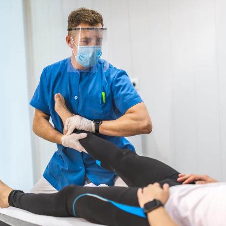 A physiotherapist with mask and screen giving a massage to an ankle. Physiotherapy with protective measures for the Coronavirus pandemic, COVID-19. Osteopathy, therapeutic chiromassage