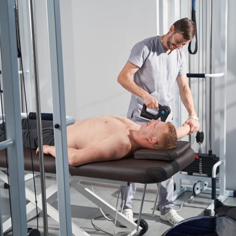 Patient lying on massage table for percussion gun treatment in rehabilitation gym. Sport injury